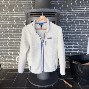 Vit fleecejacka  med blå kontraster från Patagonia - Säljer en vit fleecejacka från Patagonia med blå detaljer och dragkedjor. Jackan har tre fickor med dragkedja, hög krage och ett mjukt, fluffigt material som håller dig varm. Perfekt för kyliga dagar och har en clean, sportig look.