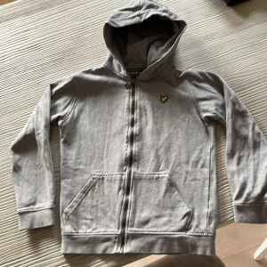 Grå hoodie från Lyle & Scott Junior - Grå hoodie från Lyle & Scott Junior med dragkedja framtill och klassisk gul broderad logga på bröstet. Tröjan har huva, känguruficka och långa ärmar. Perfekt för chill dagar eller när du vill ha en enkel och stilren look. Pris kan diskuteras
