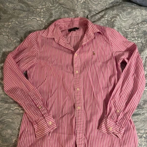 Rosa randig skjorta från Ralph Lauren - Snygg rosa och vit randig skjorta från Ralph Lauren med klassisk krage och broderad logga på bröstet. Custom fit-modell med långa ärmar och knappar vid manschetterna. Perfekt för dig som gillar stilrena och ikoniska plagg.