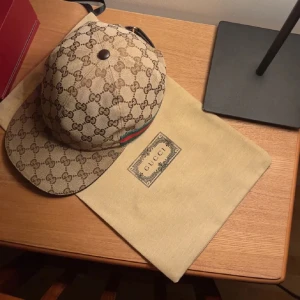 Gucci keps med GG-mönster och läderdetalj - Säljer en beige och brun keps från Gucci med klassiskt GG-monogram över hela kepsen. Kepsen har justerbar rem i brunt läder baktill och en liten grön-röd rand på sidan. Tillverkad i Italien och levereras med originalpåse.