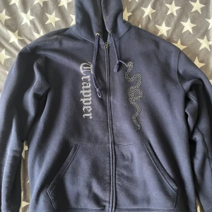 Made by trappers hoodie - Snygg mörkblå hoodie från Trapper med strassdetaljer i form av en orm och logotyp på framsidan. Tröjan har huva med snörning, dragkedja och två stora fickor framtill. Insidan är mjuk och fleecefodrad för extra komfort.