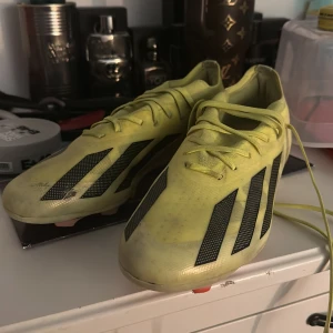 Gula Adidas fotbollsskor med svarta ränder - Säljer ett par gula Adidas fotbollsskor med klassiska svarta ränder på sidorna. Skorna har snörning och är tillverkade i syntetmaterial för lätt känsla och bra grepp på planen. Perfekta för dig som vill sticka ut på fotbollsplanen.