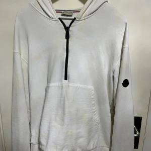 Vit hoodie från Moncler med half zip - Vit hoodie från Moncler med svart half zip och stor ficka framtill. Huva med Moncler-logga tryckt i svart och patch på ärmen. Mjuk bomullskvalitet och klassisk passform, perfekt för chill dagar. Snygg detalj med Moncler-märke bak på ryggen.