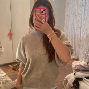 Beige stickad tröja från VILA - Supermysig beige stickad tröja från VILA med ballongärmar och ribbad hög hals. Tröjan har loose passform och breda muddar vid ärmslut och nederkant. Perfekt för lager-på-lager och chill dagar. Snygg att styla med jeans eller kjol.