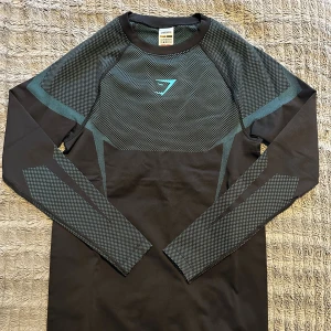 Onyx Gymshark m - Snygg svart långärmad träningströja från Gymshark med grått mönster och turkos logga på bröstet. Tröjan har en slim passform och är tillverkad i ett stretchigt syntetmaterial som andas, perfekt för gymmet eller sport. Stilren design med diskreta detaljer på ärmar och axlar.