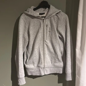 Grå hoodie från Sail Racing med dragkedja - Snygg ljusgrå hoodie från Sail Racing med hel dragkedja framtill och extra dragkedjeficka på bröstet. Tröjan har broderad logga, huva och ribbade muddar. Perfekt för chill dagar eller när du vill ha en sportig look.