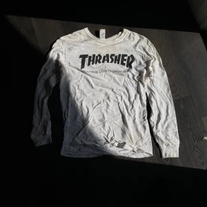 Vit långärmad Thrasher-tröja - Vit långärmad tröja från Thrasher med klassisk svart logga på bröstet. Tröjan har rund halsringning och är tillverkad i mjuk bomull, perfekt för en avslappnad streetwear-look.
