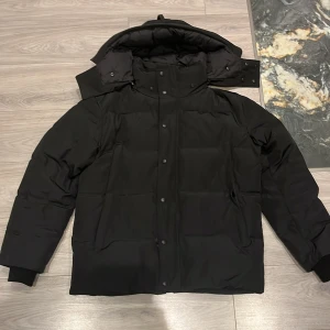 Svart pufferjacka Canada Goose  - Svart pufferjacka från Canada Goose i storlek XL. Jackan har en hög krage, avtagbar huva och knäppning med både dragkedja och knappar. Fodrad för extra värme och har ribbade muddar vid ärmslut. Perfekt för kalla vinterdagar.