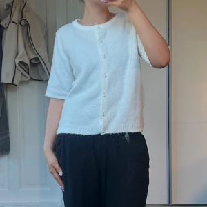 Vit kortärmad stickad tröja  från 157 - Supersöt vit blus från 157 med korta ärmar och rund hals. Blusen har pärlknappar framtill och är stickad i ett mjukt material. Perfekt för dig som gillar en clean och enkel stil med en touch av elegans.