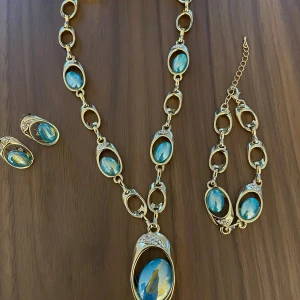 Guldförgyllt smyckeset med blå stenar - Snyggt smyckeset med halsband, armband och örhängen. Ovala länkar med blanka blå stenar och små glittriga detaljer. Stenarna har en cool marmorering i blått och gult som ger en unik vibe. Perfekt för dig som gillar statement-smycken och färg.