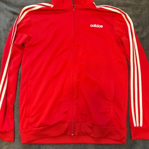 Röd Adidas vindjacka med vita ränder - Säljer en röd Adidas vindjacka i storlek L med klassiska vita ränder längs ärmarna och Adidas-logga på bröstet. Jackan har hel dragkedja framtill, ribbade muddar och två sidofickor. Perfekt för dig som gillar sportig stil och streetwear.