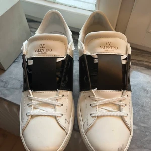 Valentino Garavani Open sneakers vit/svart - Säljer ett par snygga Valentino Garavani Open sneakers i vitt skinn med bred svart läderdetalj över snörningen. Klassisk rund tå, vita skosnören och diskret logga på plösen. Kommer med originalkartong och dustbags. Perfekta för dig som gillar exklusiva och stilrena sneakers.