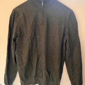 Mörkgrön stickad half zip från La Chemise - Mörkgrön stickad tröja från La Chemise med half zip-dragkedja och hög krage. Tröjan har långärmad passform och ribbade muddar vid ärmslut och nederkant. Perfekt för lager-på-lager under kyliga dagar.