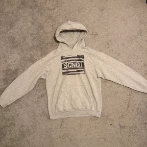 SCNDI Hoodie - Säljer en ljusgrå hoodie med svartvitt SCNDI-tryck på bröstet. Tröjan har huva, känguruficka och långa ärmar. Tillverkad i en mjuk bomulls- och polyestermix, perfekt för chill dagar. Storlek M