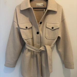 Beige kappa - Snygg beige kappa från NLY Trend med stora bröstfickor, knappar framtill och ett matchande bälte i midjan. Jackan har klassisk krage och långa ärmar med knappdetalj vid mudden. Perfekt för lager-på-lager och chill stil. Väl använd och har några små fläckar som säkert går bort vid tvätt. Den är oversized och passas XS-M