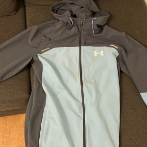 Grå och ljusblå vindjacka Under Armour - Snygg vindjacka från Under Armour i grått och ljusblått med huva och hel dragkedja framtill. Jackan har sportig design, två sidofickor och logga på bröstet. Perfekt för dig som gillar en clean och modern look. Dm för fler bilder 