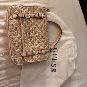 Quiltad axelväska från Guess i tweed - Superfin axelväska från Guess i beige och ljusrosa tweed med quiltad struktur och glittriga detaljer. Väskan har snygga spännen, kedjerem och flera fack inuti. Perfekt för dig som vill ha en chic och trendig look.