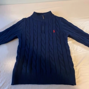 Marinblå stickad tröja Polo Ralph Lauren - Säljer en marinblå kabelstickad tröja från Polo Ralph Lauren med halv dragkedja och röd broderad logga på bröstet. Tröjan har hög krage, långa ärmar och är gjord i mjuk bomull. Perfekt för dig som gillar klassisk och stilren look.