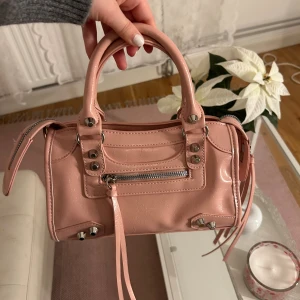 Rosa handväska med silverdetaljer - Söt rosa handväska i glansigt skinnliknande material med silverfärgade nitar och dragkedja framtill. Väskan har två handtag och dekorativa remmar samt en innerficka. 