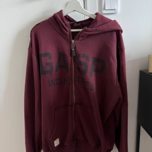Vinröd hoodie från GASP Industries - Säljer en vinröd hoodie från GASP Industries med svart tryck framtill. Tröjan har dragkedja, huva och stora fickor framtill. Perfekt för dig som gillar en sportig och avslappnad stil.
