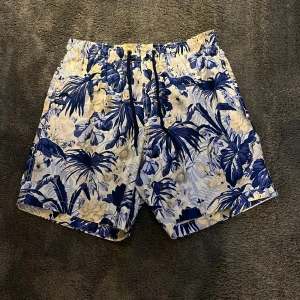 Badshorts  - Säljer ett par snygga badshorts från H&M i storlek S. De har ett blått och beige blommönster på vit botten, elastisk midja med svart snörning och innerbyxa i mesh. Perfekta för stranden eller poolen!