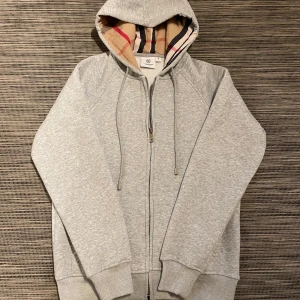 Grå hoodie från Burberry med dragkedja och kvitto - Säljer en burberry hoodie i mycket bra skick! Tröjan är köpt på NK i Göteborg när jag va där för 3799kr. Digitalt kvitto finns. Inuti tröjan så är det ett mjukt och fint material som håller dig extra varm nu när de börjar bli kyligare dagar! Om ni har några frågor så är de bara att höra av er!😊