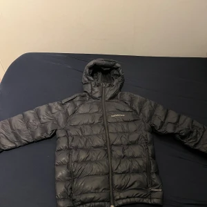 Vinterjacka Peak Performance - Säljer en svart dunjacka från Peak Performance med huva och dragkedja framtill. Jackan har quiltad design och två sidofickor med dragkedja. Perfekt för kalla dagar och har en clean look som funkar till det mesta.
