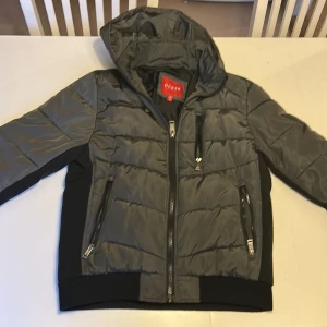 Grå pufferjacka från Guess med huva - Snygg grå pufferjacka från Guess med svart panel på sidorna och huva. Jackan har tre framfickor med dragkedja, quiltad design och ribbad nederkant. Perfekt för dig som vill ha en stilren och varm jacka med coola detaljer. Håller värmen ganska bra har bara använt den som högst 3 dagar. Har inga hål 