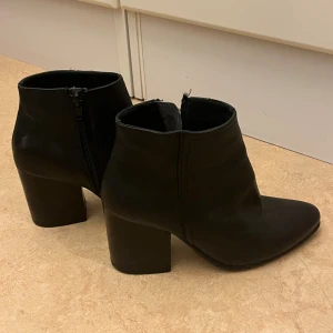Svarta stövletter från KC Cobler - Säljer ett par svarta stövletter från KC Cobler i skinn med spetsig tå och bred hög klack. Skorna har dragkedja på insidan för enkel påtagning och en stilren, minimalistisk look som passar till många outfits.