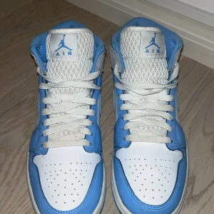 Nike Air Jordan 1 Mid UNC blå/vit - Säljer ett par Nike Air Jordan 1 Mid i UNC-färg. Skorna har en snygg mix av ljusblått och vitt skinn med grå mockadetaljer och klassisk Jordan-logga på sidan. Perforerad tå, snörning och ikonisk swoosh. Perfekta för dig som vill sticka ut med stil.