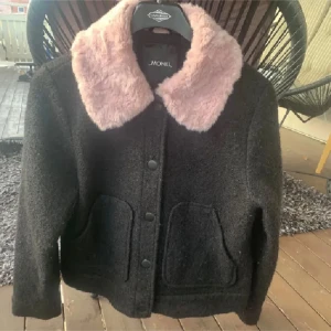 Svart jacka med rosa fuskpäls Monki - Svart jacka från Monki med stor fluffig krage i ljusrosa fuskpäls. Jackan har två rejäla fickor framtill, knäppning med svarta knappar och en boxig, loose passform. Materialet är grov ullmix som ger en cool struktur. Perfekt statementkrage för dig som gillar att sticka ut. Kan va lite nopprig
