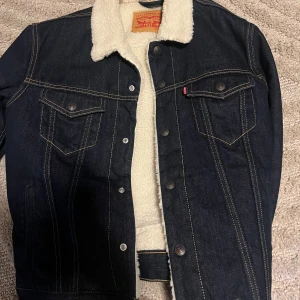 Levi's jeansjacka med teddyfoder M - Säljer en mörkblå jeansjacka från Levi's med mjukt vitt teddyfoder och krage. Jackan har klassiska bröstfickor med knappar, kontrastsömmar och en schysst vintage-vibe. Perfekt för höst och vinter när du vill vara både varm och snygg.
