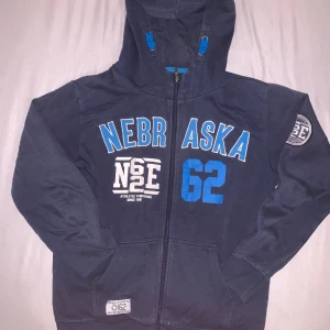 Mörkblå hoodie Nebraska Red & Blue - Mörkblå hoodie från Red & Blue med Nebraska-tryck och siffran 62 i blått framtill. Tröjan har huva med blå detaljer, dragkedja och ribbade muddar. Patch med NSE på ena ärmen och fickor framtill. Perfekt för dig som gillar sportig college-stil.