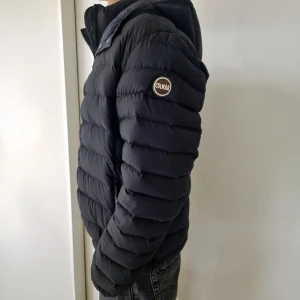 Svart Colmar pufferjacka med huva - Svart pufferjacka från Colmar med huva och dragkedja framtill. Jackan har ett quiltat mönster och logotyp på vänster ärm. Passformen är normal och jackan är perfekt för kyliga dagar. Snygg och stilren design som funkar till jeans och sneakers.