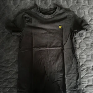 Svart t-shirt från Lyle & Scott - Svart t-shirt från Lyle & Scott med klassisk passform och rund hals. På bröstet sitter den ikoniska gula örnloggan. Tillverkad i mjuk bomull, perfekt för en clean och stilren look.