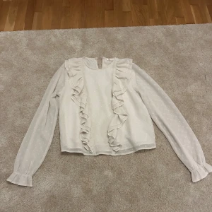 Vit blus med volanger från Gina Tricot - Supersöt vit blus från Gina Tricot med diskret prickigt mönster och långa ärmar. Blusen har volanger framtill och vid ärmslut, samt en liten knapp i nacken. Perfekt för dig som gillar romantisk och feminin stil!