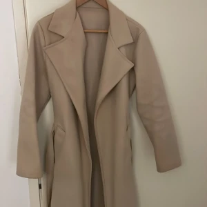 Beige lång kappa  - Snygg beige kappa som har långa ärmar och två fickor framtill. Storlek S passar även Xs. Perfekt till våren/höst 