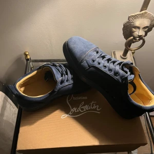 Blå sneakers från Christian Louboutin - Snygga blå sneakers från Christian Louboutin i mocka med klassisk röd sula och svarta snören. Skorna har rund tå och platt sula, samt beige innersida. Perfekta för dig som vill sticka ut med exklusiva detaljer och ikonisk design.