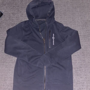 Mörkblå hoodie från Sail Racing - Mörkblå hoodie från Sail Racing med huva och dragkedja hela vägen. Snyggt broderad logga på bröstet och huvan, samt en praktisk bröstficka med dragkedja. Mjuk insida och klassisk passform, perfekt för chill dagar.