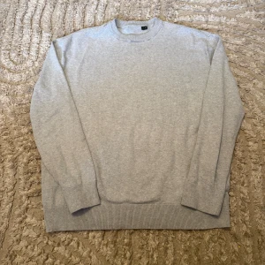 Uniqlo Grå stickad tröja - En enkel och stilren grå stickad tröja med rund hals och ribbade muddar vid ärmslut och nederkant. Perfekt för lager-på-lager och chill dagar. Tröjan har en normal passform och är långärmad, vilket gör den skön att bära under höst och vinter.