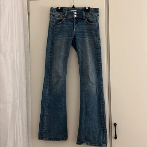 Blå bootcut jeans från Gina Tricot - Snygga blå jeans från Gina Tricot med bootcut passform och låg midja. Jeansen har två framfickor, dubbla knappar i midjan och coola bakfickor med lock och knapp. Materialet är klassisk jeans med slitningar för en trendig look.