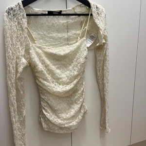 Vit spetsblus från BikBok - Säljer en superfin vit blus från BikBok i spets med långa ärmar och draperad front. Blusen har smala axelband och är genombruten med vackert spetsmönster. Perfekt för dig som vill ha en romantisk och trendig look.
