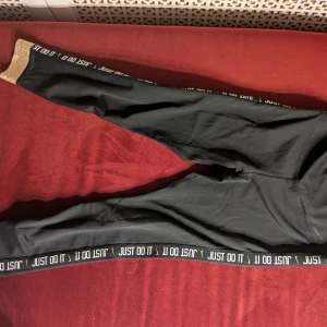 Nike Dri-Fit svarta leggings M - Svarta Nike Dri-Fit leggings med texten 'JUST DO IT' längs benen och en liten gulddetalj. Guldglittrig kant vid benslut och diskret Nike-logga på höften. Snygg och sportig look, perfekt för gymmet eller chill. Stretchigt material som sitter tight.