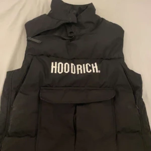  Hoodrich väst - Svart dunväst från Hoodrich med hög krage och stor ficka framtill. Västen har vit logotyp på bröstet och dragkedjor på sidorna. Perfekt för lager-på-lager och streetwear-stil.
