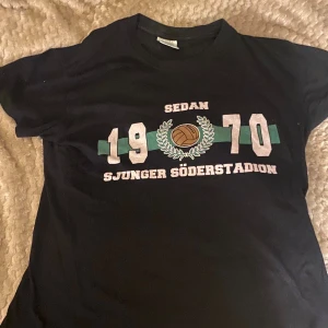 Svart t-shirt med Söderstadion-tryck - Svart t-shirt med tryck 'Sedan 1970 Sjunger Söderstadion' och en fotboll omgiven av lagerkrans. Snyggt retroinspirerat motiv i vitt, grönt och brunt. Klassisk rund hals och korta ärmar, perfekt för dig som gillar fotboll och supporterstil.