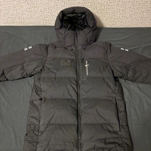 Svart dunjacka från Sail Racing - Säljer en svart dunjacka från Sail Racing med huva och hel dragkedja. Jackan är lång, har diskreta loggor på bröstet och ärmarna samt reflekterande detaljer. Perfekt för kalla dagar och riktigt snygg streetstil. Passar perfekt till folk som har storlek S-M. Priset kan sänkas vid snabb affär.