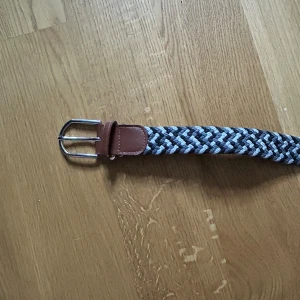 Randigt flätat bälte med skinn-detaljer - Snyggt flätat bälte med randigt mönster i svart, grått och vitt. Bältet har bruna skinn-detaljer vid spännet och änden, samt ett silvrigt metallspänne. Perfekt för att ge outfiten lite extra edge och passar till både jeans och chinos.