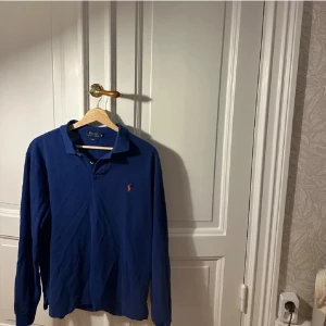 Blå långärmad pikétröja Polo Ralph Lauren - Säljer en blå långärmad pikétröja från Polo Ralph Lauren i slim fit. Tröjan har klassisk krage, knappar vid halsen och den ikoniska broderade loggan i rött på bröstet. Perfekt för dig som gillar stilren och sportig look. Passar mer som M