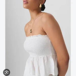 Vit bandeau tubtopp från Gina Tricot - Supersöt vit bandeau tubtopp från Gina Tricot med smockad överdel och volangkant upptill. Toppen har en luftig, rynkad nederdel som ger en flowy känsla. Perfekt för varma dagar och enkel att styla med smycken eller jeans.