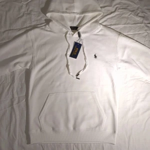 Vit hoodie från Polo Ralph Lauren - Snygg vit hoodie från Polo Ralph Lauren med klassisk broderad logga på bröstet. Tröjan har huva med snörning, känguruficka och ribbade muddar. Perfekt för en clean och stilren look. Tillverkad i mjuk bomull för maximal komfort.